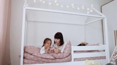 Küçük beyaz bir kız ve annesi evde akıllı telefondan müzik dinliyor, rahat bir yatak odasında rahat bir şekilde yatarken pijama giyiyor, annesini ve tatlı kızını seviyor, kulaklıklarını takıyor, kapatıyorlar.