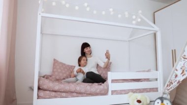 Genç anne ve kızı yatak odasında oynuyorlar. Komik anne ve güzel çocuk ahşap yatakta telefonla görüntülü görüşme yapıyorlar..