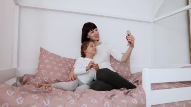 快乐的妈妈和十几岁的女儿坐在床上 在家里用智能手机应用程序大笑 快乐的积极的家庭成人父母 与十几岁的女孩一起欣赏有趣的视频 享受放松的生活 — 图库视频影像