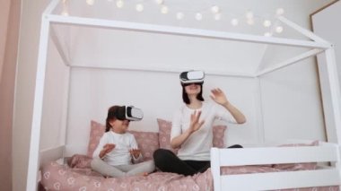 Yeni dijital teknoloji. Evde İskandinav ahşap yatağında oturmuş sanal gerçeklik gözlüğü takıp gülümseyen neşeli beyaz anne ve kız..