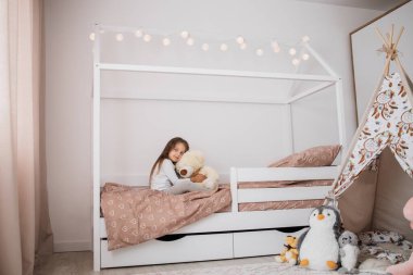 Pijama giymiş sevimli küçük bir kız İskandinav ahşap evde oyuncak ayısına sarılıyor evde, mutlu bir çocukluk konsepti, kapalı yatay portresi.