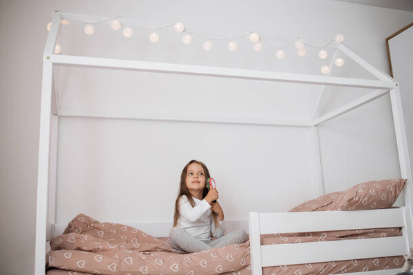 Close up portrait of sweet little girl brushing her long hair and smiling while sitting on her scandinavian wooden house frame bed at home.