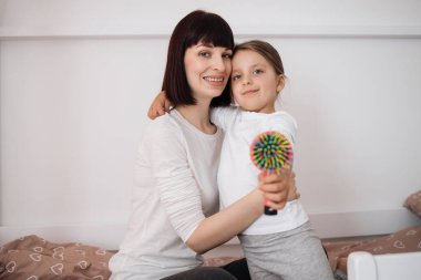 Saçını taradıktan sonra anaokulu kızını kucaklayan, yatak odasının iç kısmında, tahta yatakta oturan, tatmin olmuş, beyaz bir bayan..