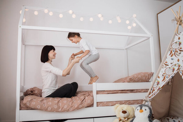 Cheerful mum babysitter play with cute active small kid girl jump on bed, happy carefree mother and little child daughter holding hands laughing having fun feel joy at home in modern bedroom interior