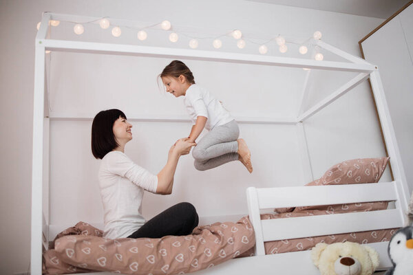 Cheerful mum babysitter play with cute active small kid girl jump on bed, happy carefree mother and little child daughter holding hands laughing having fun feel joy at home in modern bedroom interior
