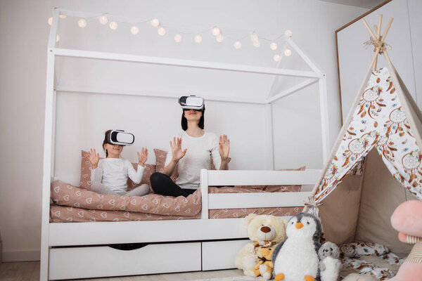 Новые цифровые технологии. Радостная милая белая мать и дочь улыбаются, сидя дома в очках виртуальной реальности на скандинавской деревянной кровати.
