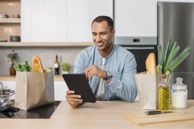 Çekici genç Hintli adam vejetaryen sebze salatası tarifini okumak için dijital tablet kullanıyor. Evde modern, parlak bir mutfakta yemek pişirirken oturup gülümsüyor..