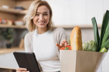 Elinde tablet ve içinde sebzeler olan kağıt torbalara odaklan. Mutlu kadın sağlıklı yemek pişiriyor sebzeli kâğıt torbaya bakıyor ve evdeki modern mutfakta dijital tablette yemek tarifi okuyor..