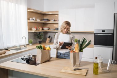 Neşeli genç, beyaz, sarışın kadın evde akşam yemeği için salata pişiriyor, dijital tablete bakıyor, yemek blogu okuyor, mutfak içini okuyor, fotokopi çekiyor. Aşçılık, aşçılık konsepti.