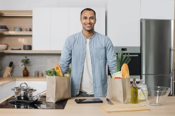 Young positive attractive indian man preparing salad, healthy food, from fresh organic vegetables, standing on the background of new modern light kitchen at home.