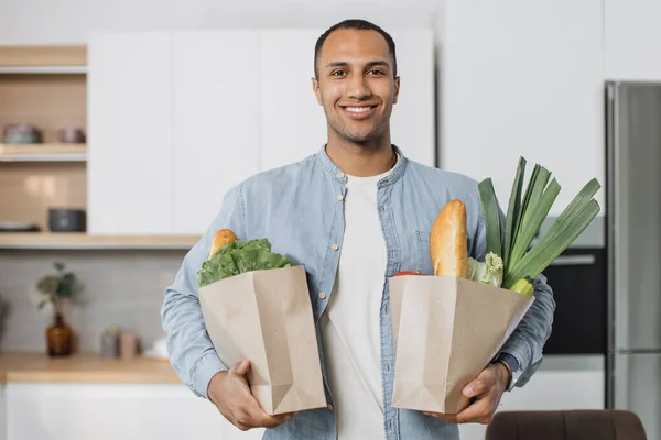 Young attractive indian man in kitchen holding two paper grocery shopping bags full of fresh healthy food vegetables in his hand, standing on the background of new modern light kitchen at home.