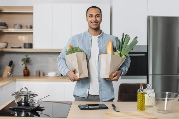 Young attractive indian man in kitchen holding two paper grocery shopping bags full of fresh healthy food vegetables in his hand, standing on the background of new modern light kitchen at home.