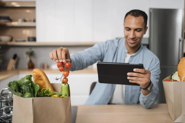 Focus on hand of blurred indian man in kitchen taking cherry tomatoes from grocery shopping bag full of fresh healthy food vegetables lying on new modern kitchen table at home.