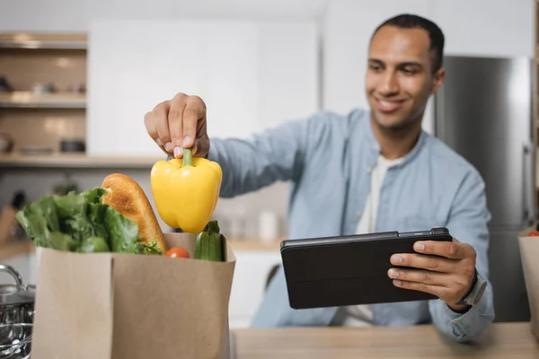 Focus on hand of blurred indian man in kitchen taking yellow pepper from grocery shopping bag full of fresh healthy food vegetables lying on new modern kitchen table at home.