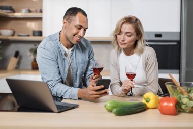 Gülümseyen genç eşler birlikte sağlıklı bir kahvaltı yapıyorlar ve cep telefonu kullanıyorlar, online alışveriş yapıyorlar ya da internette sörf yapıyorlar, mutlu bir aile yeni mutfak ortamında sebze yiyor..