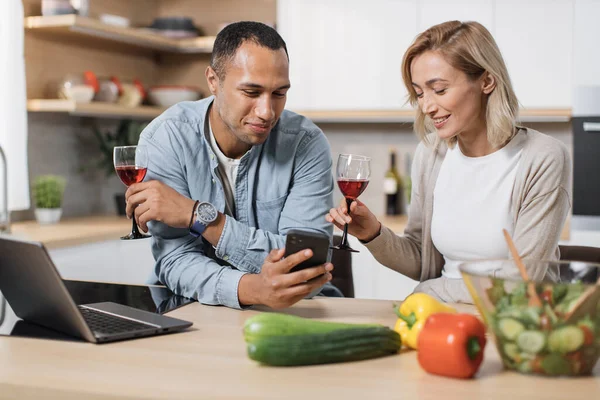 Distracted from cooking happy young multinational family couple using cellphone checking interesting food recipe drinking wine communicating online inviting friends for party preparing meal in kitchen
