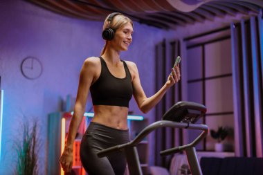 Aktif ve dinamik kulaklıklı genç bir kadın evde spor yapıyor. Koşu bandında geceleri cep telefonuyla koşuyor. Vücudunu düzelt. Mükemmel durumda. Dayanıklılık artırılıyor.