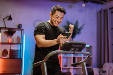 Heyecanlı genç Asyalı sporcu, spor yaparken, spor yaparken, koşu bandında koşarken akıllı telefona bakarak iyi sonuçlar elde ediyor. Atletik ve kaslı erkek aktif olarak kapalı alanda egzersiz yapıyor..