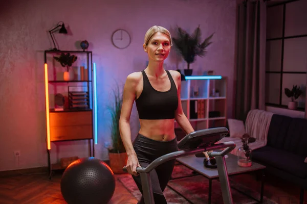 Side view of blond beautiful woman in sports clothes doing cardio training on treadmill at night at home or gym. Concept of sport, health care, action, remote leisure.