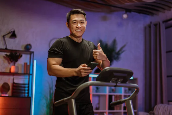 Portrait of active asian man in sportswear using smartphone, running using treadmill at home at night. Coronavirus Covid 19 social distance. Home workout, sports, stay at home, remote leisure concept.