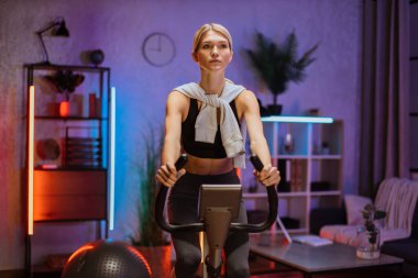 Spor sutyen giyen ve egzersiz bisikleti kullanan odaklanmış genç bir kadının portresi. Akşamları evde spor yapan sporcu bir kadın akıllı staj bisikleti üzerinde çalışıyor. Genç beyaz atlet kız..