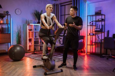 Spor kıyafetleri içinde güzel sarışın beyaz spor kadınlar bisiklet sürüp akşamları spor eğitmeninin talimatlarını dinliyorlar. Kardiyo eğitimi, egzersiz bacakları, içeride kardiyo çalışması..