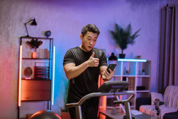 Side view of young confident handsome asian man looking at smartphone during video call while running on treadmill at modern apartment or gym. Jogging with pleasure.