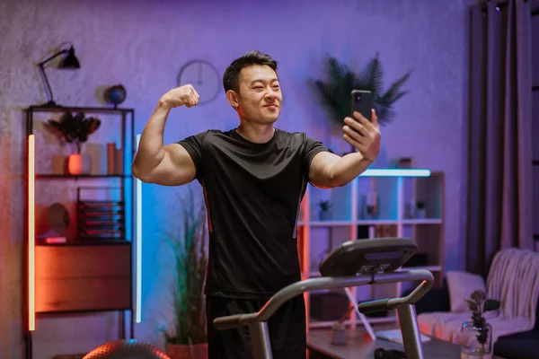 Concept of sports training in evening time at home. Young attractive asian man showing arm muscles, bicep while looking at smartphone, doing selfie during exercising on treadmill.