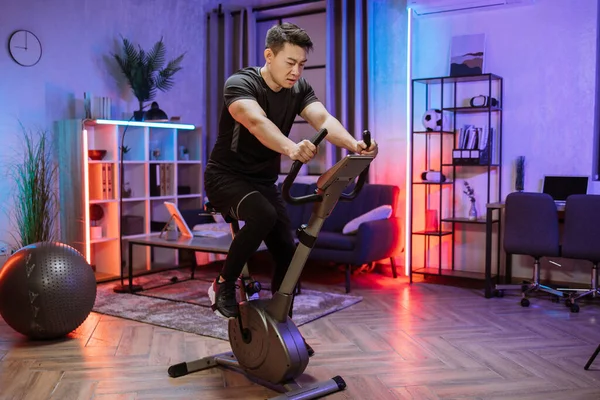 Home fitness workout, young asian man athlete training on smart stationary bike indoors watching on screen connected online to live streaming subscription service for biking exercise.