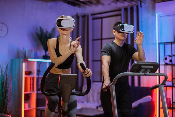 Home fitness workout sporty people caucasian woman and asian man training on exercise machines, using 3d virtual glasses imitating outdoor activities, at evening time at home.