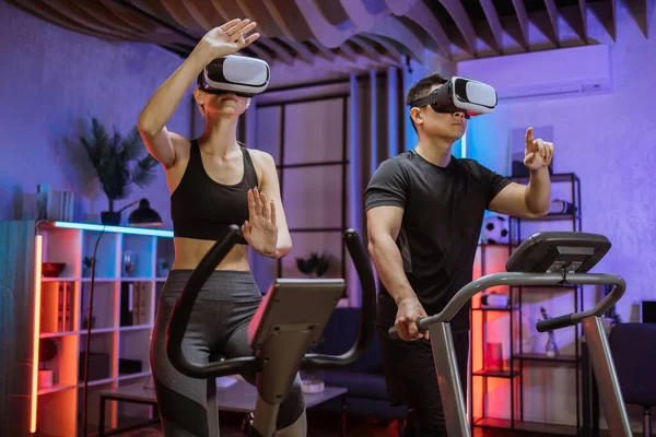 Joyful caucasian woman training on exercise bike with sporty asian man running on treadmill and using 3d virtual reality glasses for playing and simulation competitions in the stadium.