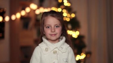 Süslü Noel ağacının önünde, kalın örülü beyaz süveter giyen ve kameraya bakan sevimli küçük kızın video portresini kapat..