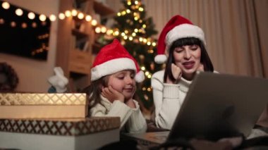 Güzel, mutlu bir genç kadın ve kızının samimi bir Noel arifesinde dizüstü bilgisayardan alışveriş yaparken çekilmiş bir fotoğrafı. Anne yerde, Noel ağacının yanında yatıyor ve kız çocuğu da yanında, alışveriş hediyeleri..