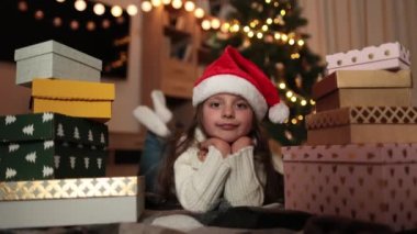 Süslü Noel ağacının önünde yere uzanmış, kalın örgü beyaz süveter ve kırmızı Noel Baba şapkası giyen sevimli küçük kızın video portresini kapat ve kameraya bak..
