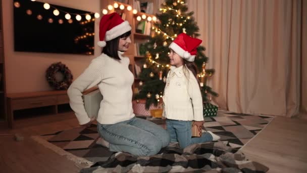 圣诞节家庭庆祝活动 年轻女子笑着 坐在装饰过的树上 和可爱的学龄前女儿戴着圣诞礼帽共享圣诞礼物 在家里度过了一个快乐的寒假 — 图库视频影像