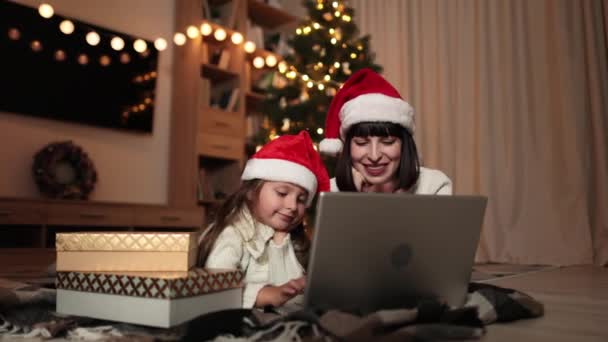 年轻的高加索人母亲和她可爱的小女儿正在用笔记本电脑与人聊天 她们躺在自家客厅的地板上 房间里装饰着圣诞树 — 图库视频影像