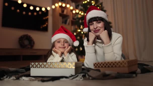 傍晚时分 漂亮的白俄罗斯年轻妈妈和她可爱的学龄前女儿躺在地板上 带着礼物躺在装饰过的房间里模糊的圣诞树前 — 图库视频影像
