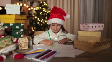 Mutlu Noeller ve mutlu tatiller. Kırmızı şapkalı şirin kız Noel Baba 'ya mektup yazıyor. Evdeki Noel ağacının yanındaki masada oturuyor..