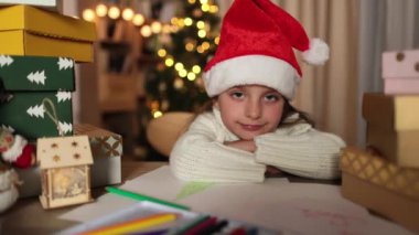 İyi kız hediyeleri bekliyor. Noel Baba şapkalı güzel bir kız mektup yazıyor ve Noel ağacının arkasındaki hediye yığınının yanında sevinçle gülümsüyor..