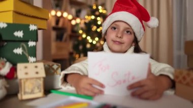 Noel, tatiller ve çocukluk konsepti. Kırmızı şapkalı, gülümseyen beyaz kız. Evde Noel Baba için dilek listesi yazdıktan sonra el ele tutuşuyor..