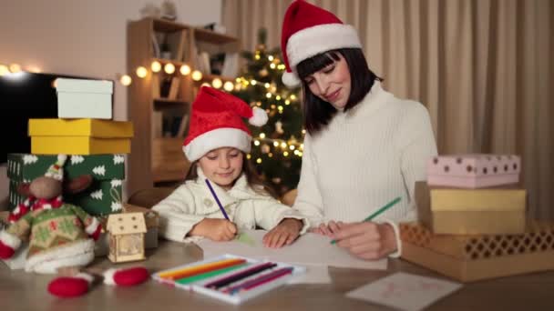 在装饰过的客厅里 学龄前小女孩坐在漂亮的母亲身旁 嘴角挂着迷人的笑容 一边写着给圣诞老人的信 一边坐在桌子旁 — 图库视频影像