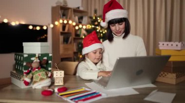 Güzel, mutlu bir genç kadın ve kızının samimi bir Noel arifesinde dizüstü bilgisayardan alışveriş yaparken çekilmiş bir fotoğrafı. Anne ve kızı Noel ağacının yanında alışveriş hediyeleriyle masada oturuyor..