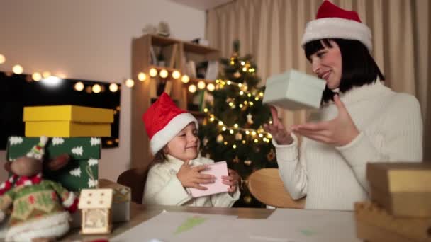 圣诞节家庭庆祝活动 年轻女子笑着和可爱的学龄前女儿戴着圣诞礼帽坐在装饰树旁的餐桌旁交换圣诞礼物 — 图库视频影像
