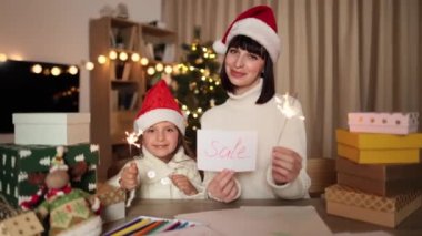 Noel Baba şapkalı sevimli kız ve annesi Noel ağacı ve dekore edilmiş oturma odasının arka planında ışıltı ve indirim kartıyla masada oturuyorlar..
