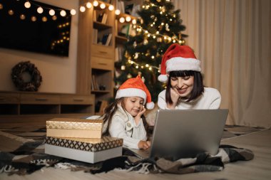 Güzel, mutlu bir genç kadın ve kızının samimi bir Noel arifesinde dizüstü bilgisayardan alışveriş yaparken çekilmiş bir fotoğrafı. Anne yerde, Noel ağacının yanında yatıyor ve kız çocuğu da yanında, alışveriş hediyeleri..
