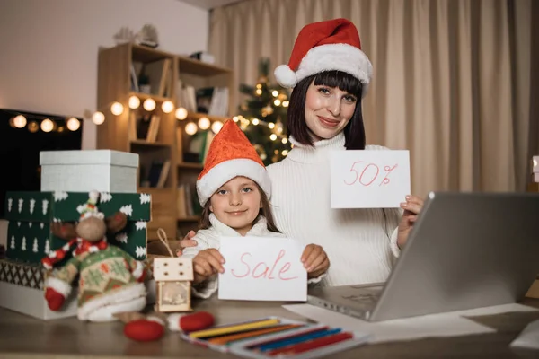Happy family, young beautiful mother and little cute child daughter in red hats holding letter with sale inscription on Christmas Eve, at evening at decorated home. Concept of holiday discount.