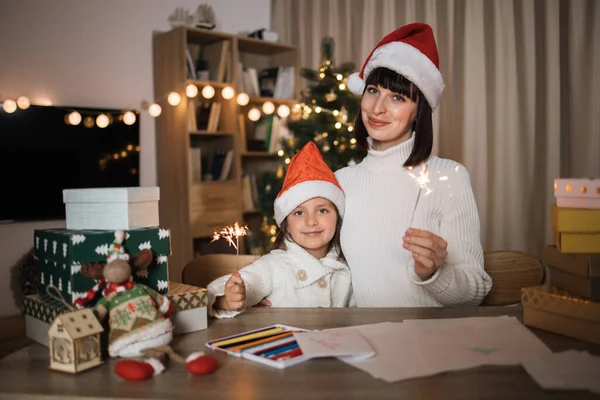 Cheerful cute girl and her mother with charming smile in Santa hats sitting on table with sparkles on the background of Christmas tree and decorated living room.