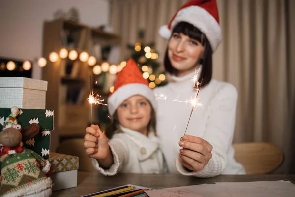 Focus on hands with sparkles, blur background of cheerful cute girl and her mother with charming smile in Santa hats sitting on table on the background of Christmas tree and decorated living room.