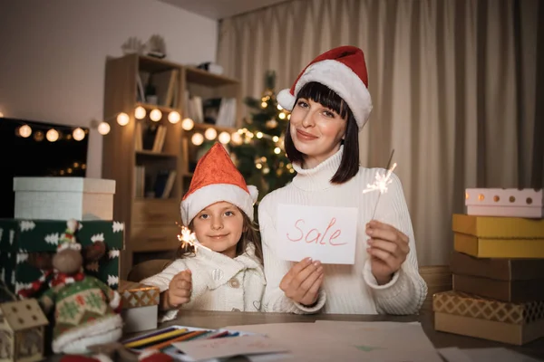 Cheerful cute girl and her mother with charming smile in Santa hats sitting on table with holiday sale discount card and sparkles on the background of Christmas tree and decorated living room.