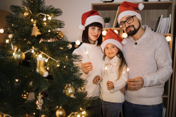 Family, winter holidays and people concept - bearded caucasian father, young mother and little cute daughter holding sparkles, bengal fire on background of decorating Christmas tree at home.